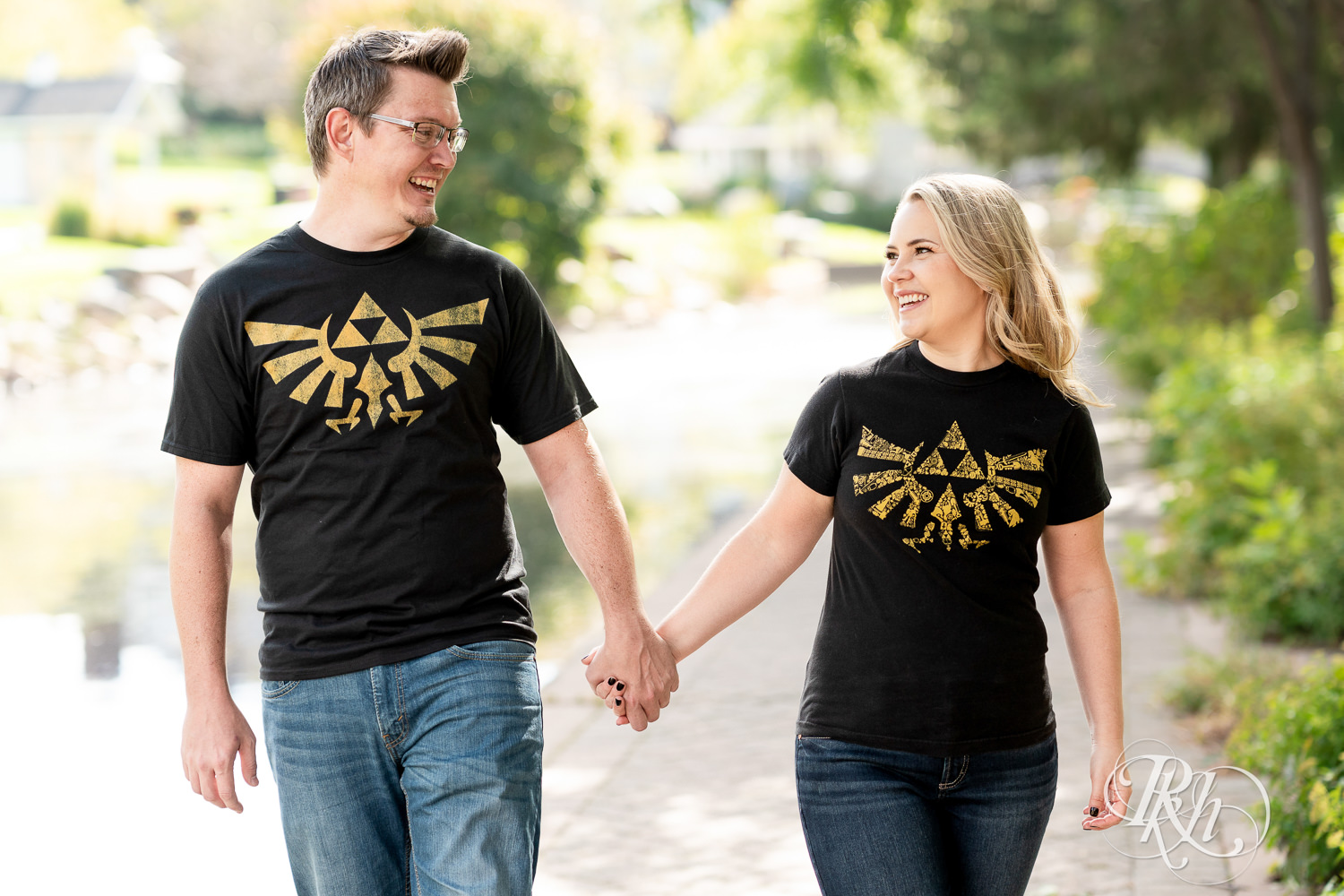 Man and woman in Zelda shirts walk along lake during engagement photography at Centennial Lakes Park in Edina, Minnesota.