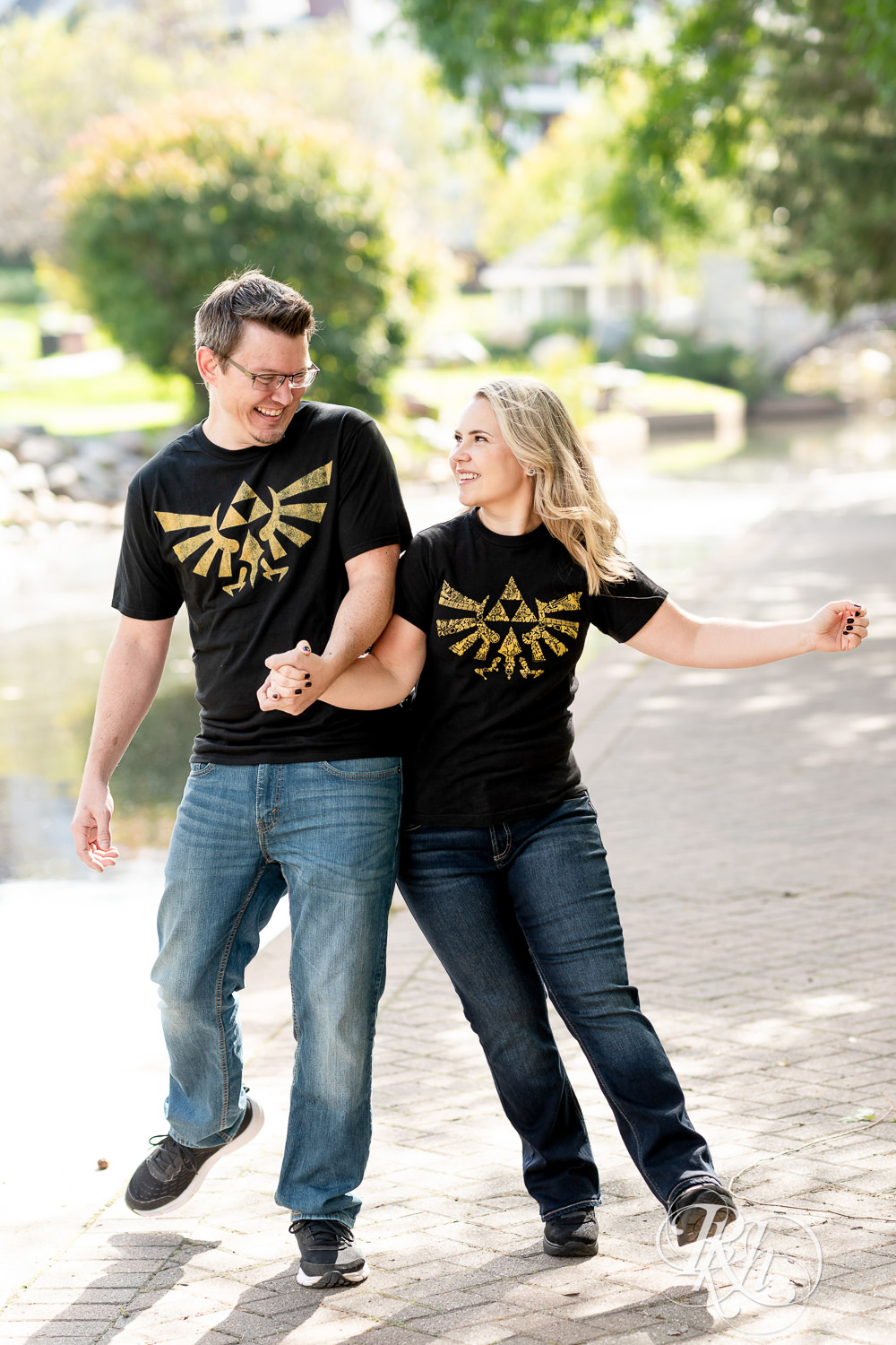 Man and woman in Zelda shirts walk along lake during engagement photography at Centennial Lakes Park in Edina, Minnesota.