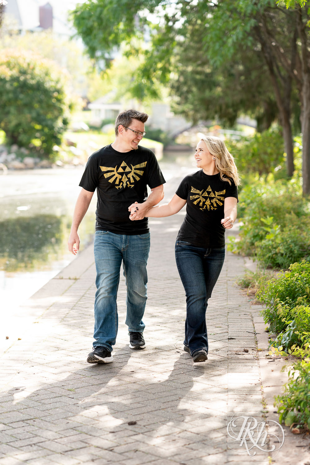 Man and woman in Zelda shirts walk along lake during engagement photography at Centennial Lakes Park in Edina, Minnesota.