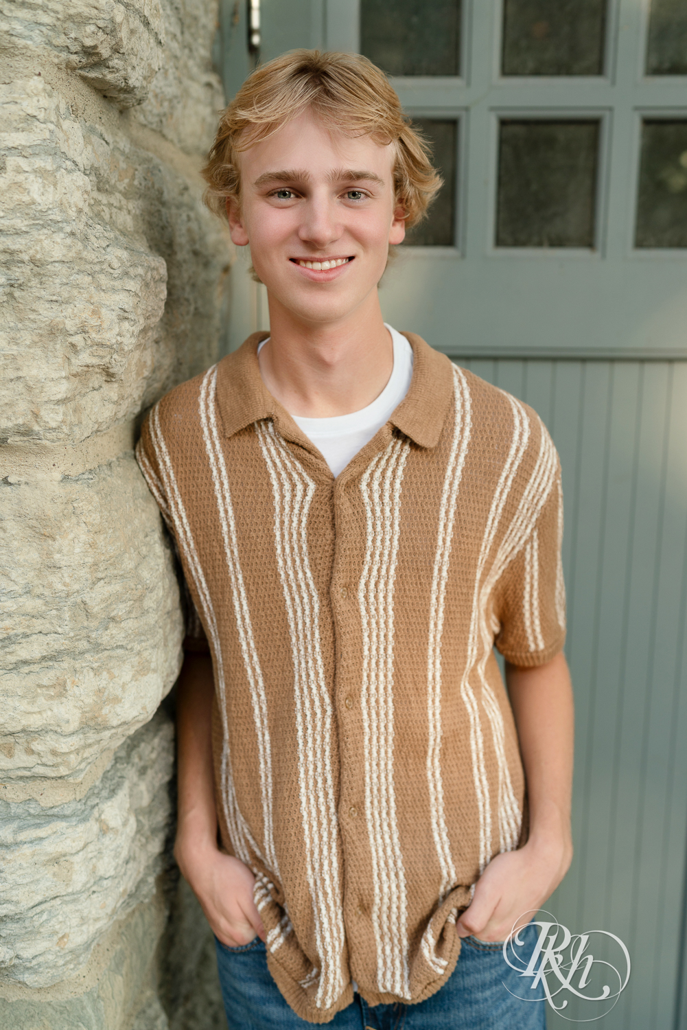 Blond senior boy dressed in jeans smiles standing against wall at sunset in Minneapolis, Minnesota.