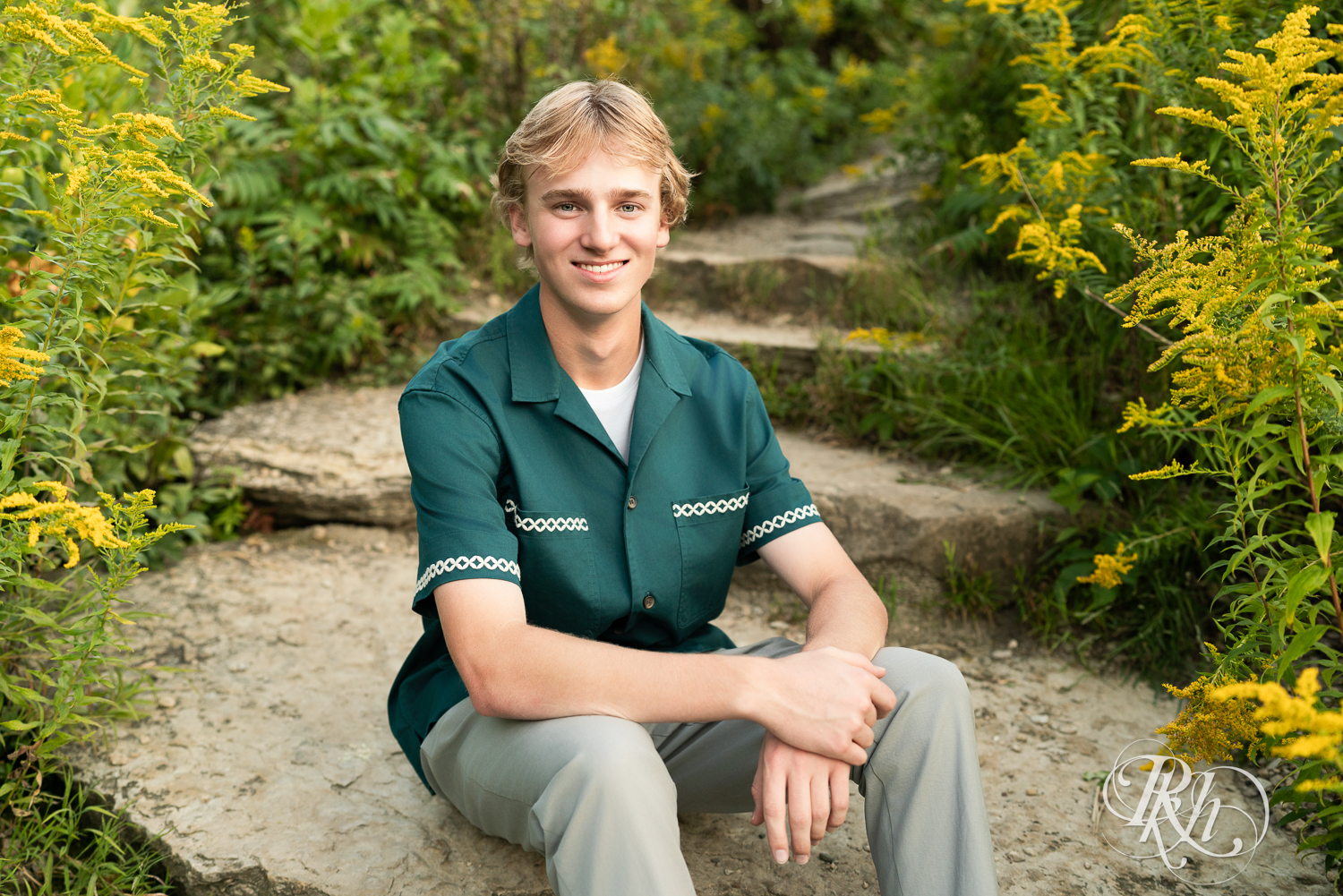 Blond senior boy dressed in green shirt and khakis smiles sitting on a rock at sunset in Minneapolis, Minnesota.