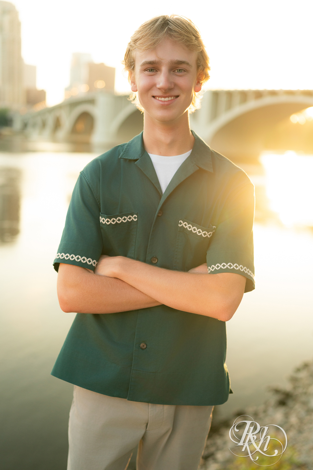 Blond senior boy dressed in green shirt and khakis smiles standing on a rock at sunset in Minneapolis, Minnesota.