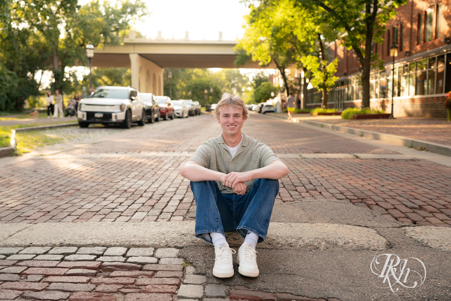 Blond senior boy dressed in jeans smiles on cobblestone street at sunset in Minneapolis, Minnesota.