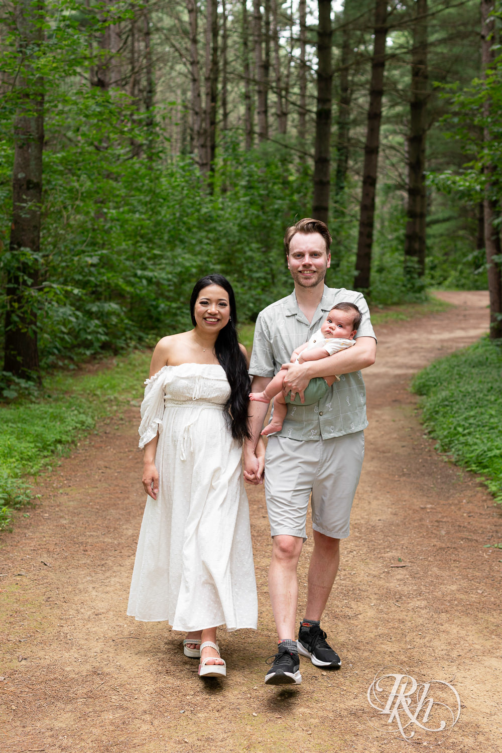 Woman in white dress and man in green dress shirt and shorts walk down a trail holding their baby at Lebanon Hills Regional Park in Eagan, Minnesota.