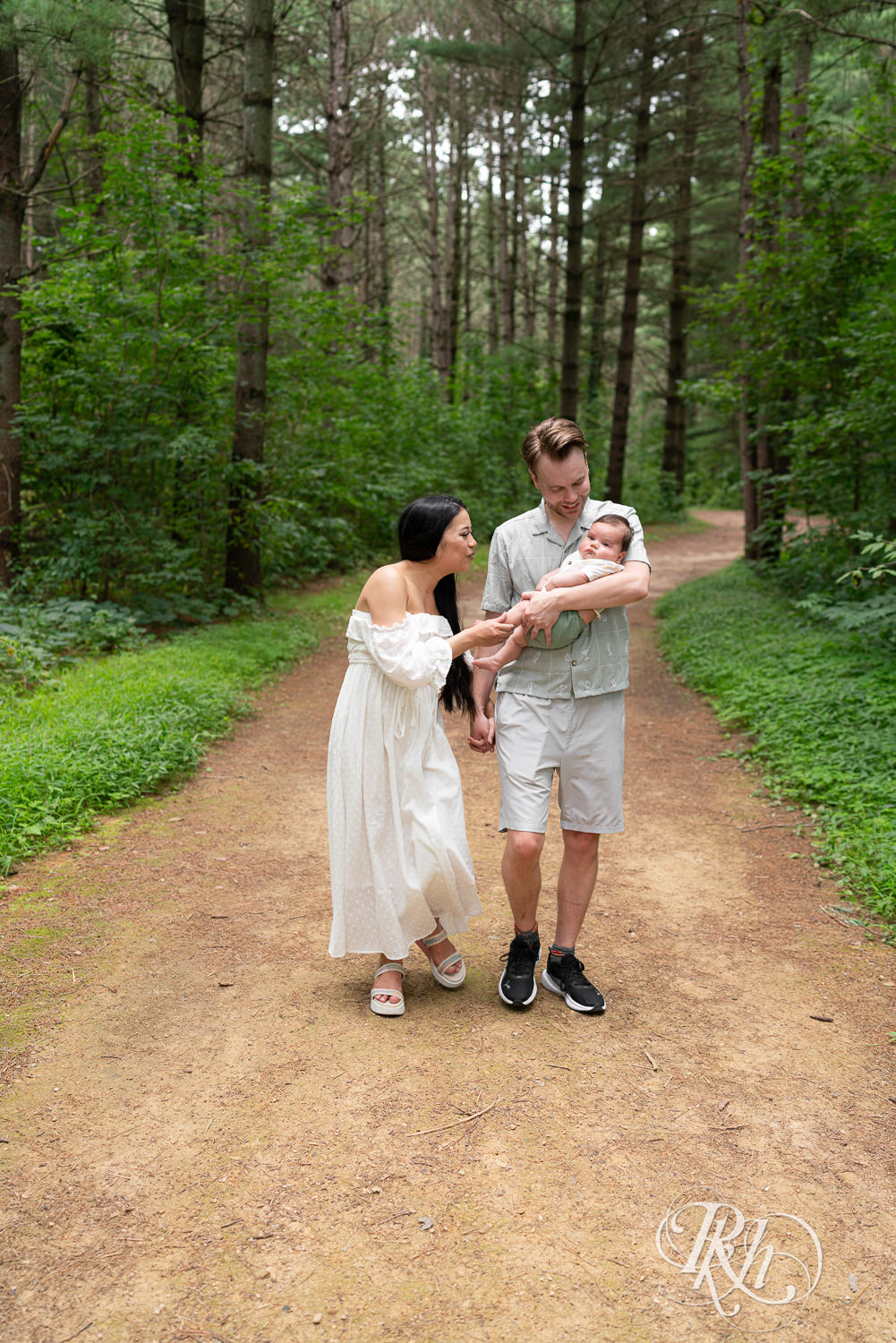 Woman in white dress and man in green dress shirt and shorts walk down a trail holding their baby at Lebanon Hills Regional Park in Eagan, Minnesota.