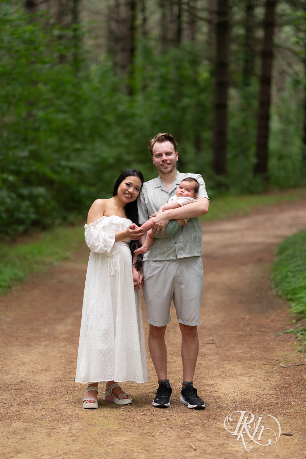 Woman in white dress and man in green dress shirt and shorts walk down a trail holding their baby at Lebanon Hills Regional Park in Eagan, Minnesota.
