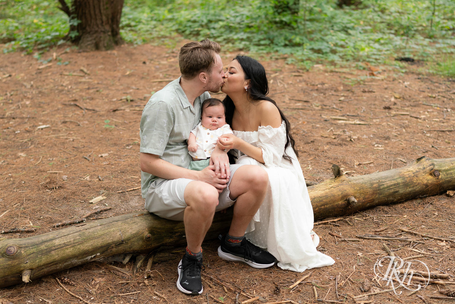Woman in white dress and man in green dress shirt and shorts kiss while holding their baby at Lebanon Hills Regional Park in Eagan, Minnesota.