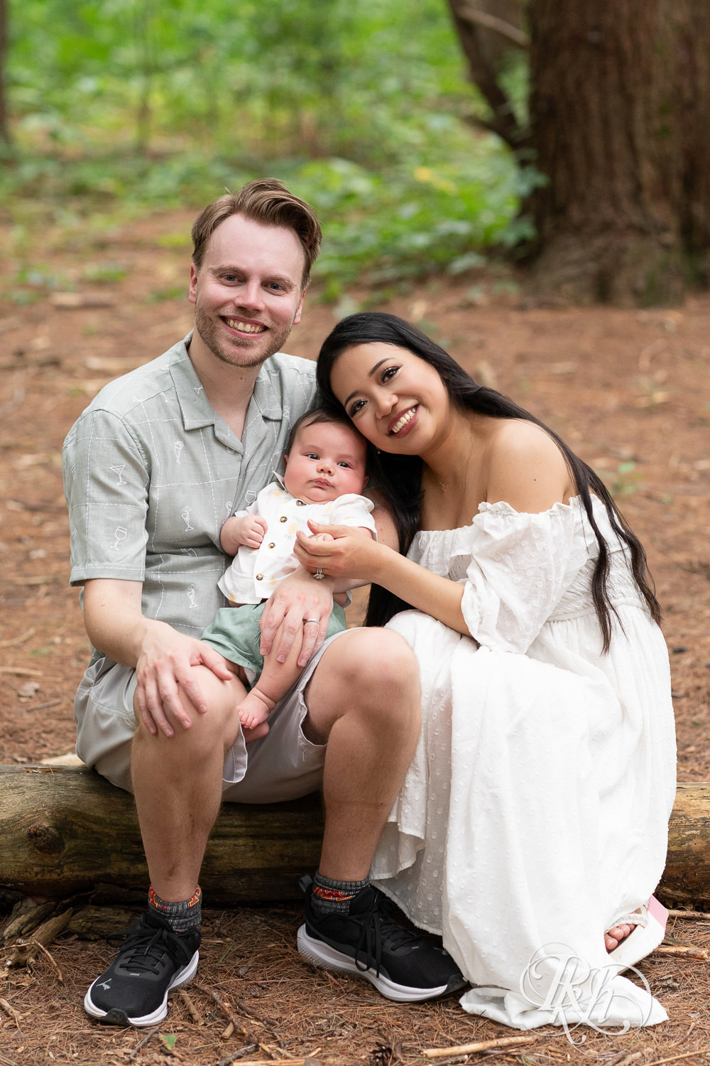 Woman in white dress and man in green dress shirt and shorts smile while holding their baby at Lebanon Hills Regional Park in Eagan, Minnesota.