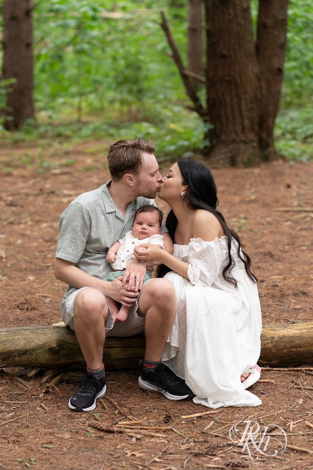 Woman in white dress and man in green dress shirt and shorts kiss while holding their baby at Lebanon Hills Regional Park in Eagan, Minnesota.