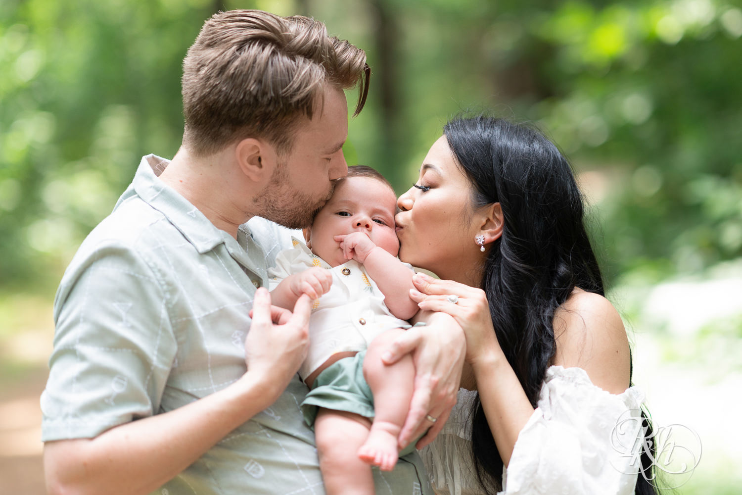 Woman in white dress and man in green dress shirt and shorts kiss their baby at Lebanon Hills Regional Park in Eagan, Minnesota.