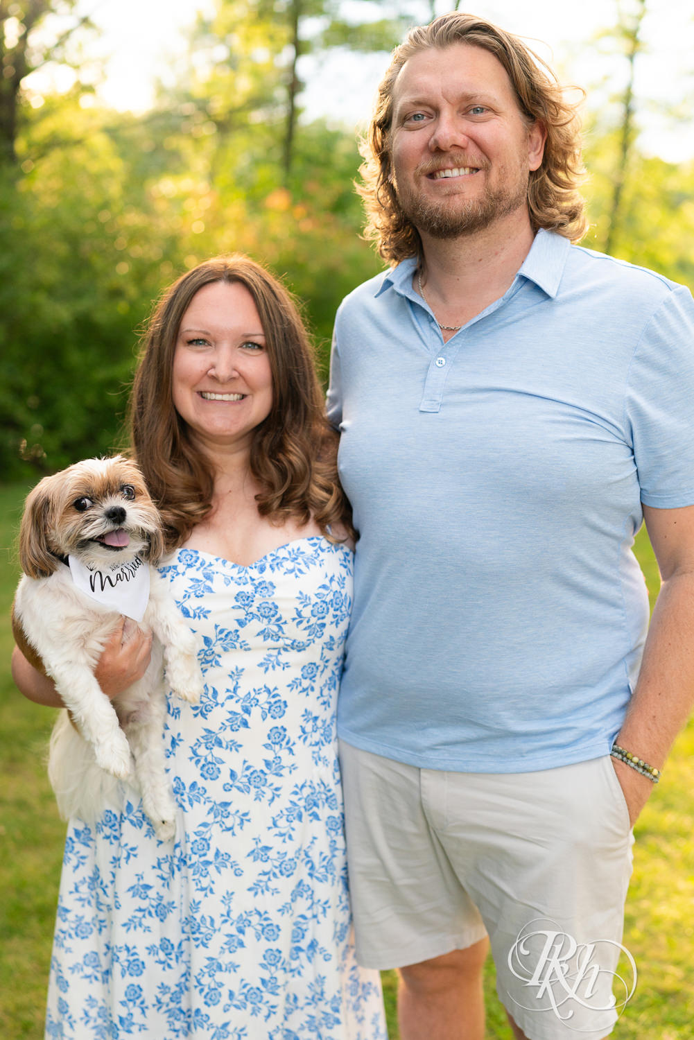 Man in blue shirt and khaki shorts and woman in blue and white dress smile while holding dog at sunset in Eagan, Minnesota.