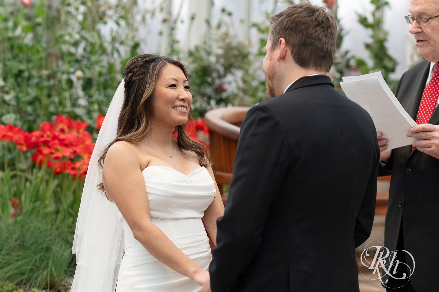 Asian bride smiles at groom during wedding ceremony in the Sunken Garden at the Como Zoo and Conservatory in Saint Paul, Minnesota.