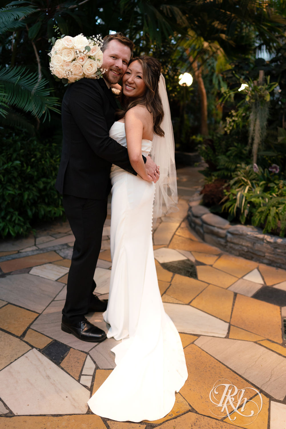 Asian bride and groom smile while hugging in the Palm Dome at the Como Zoo and Conservatory in Saint Paul, Minnesota.