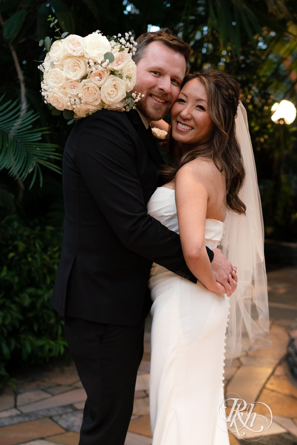 Asian bride and groom smile while hugging in the Palm Dome at the Como Zoo and Conservatory in Saint Paul, Minnesota.