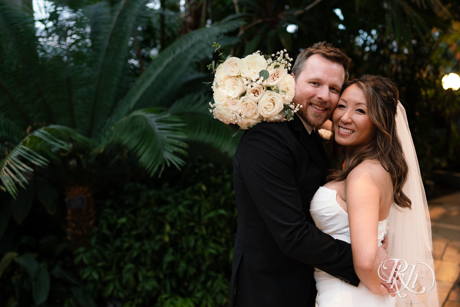 Asian bride and groom smile while hugging in the Palm Dome at the Como Zoo and Conservatory in Saint Paul, Minnesota.