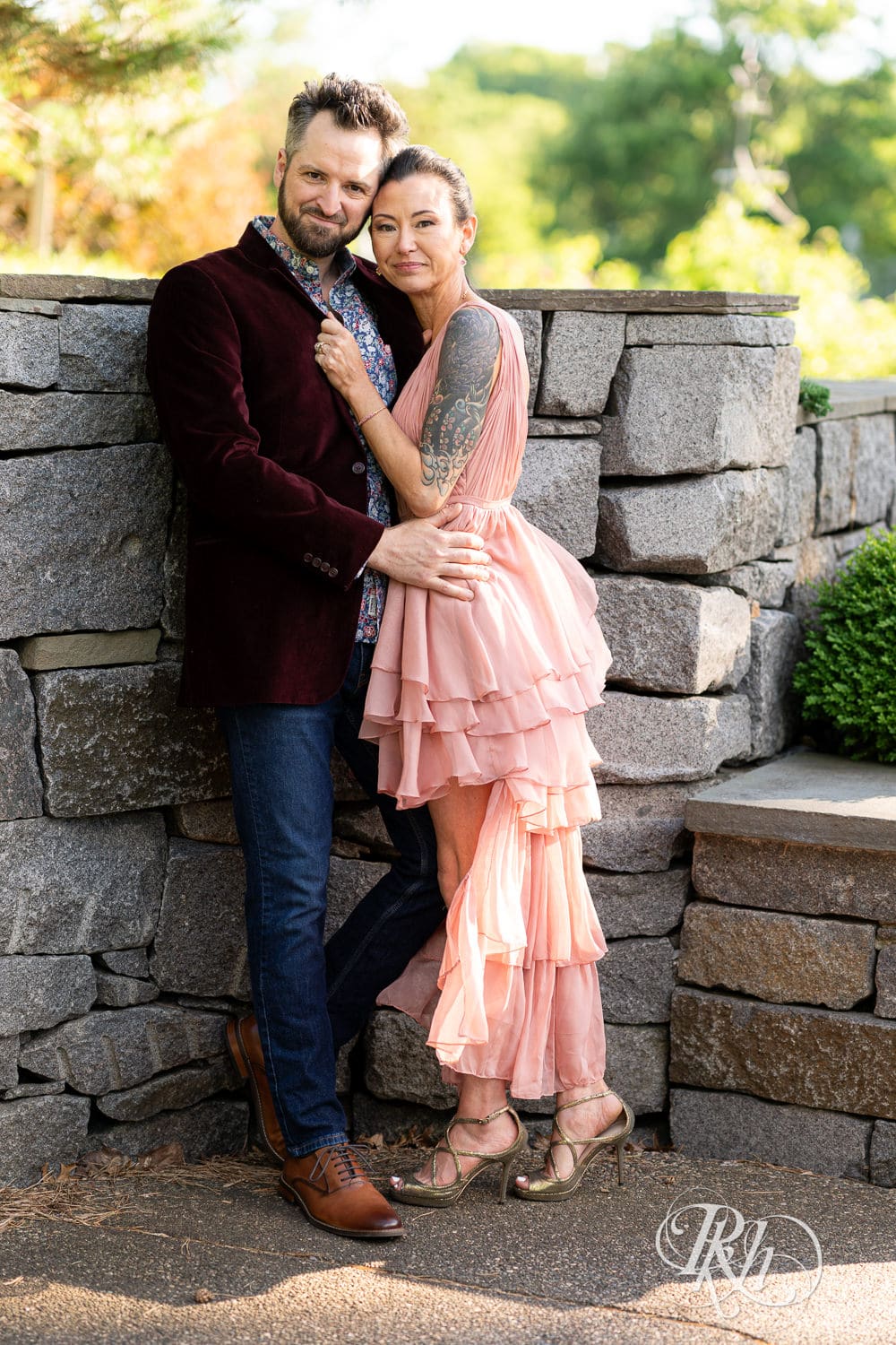 Man in red velvet jacket and a woman in a pink dress smile in front of a stone wall at the Minnesota Landscape Arboretum in Chaska, Minnesota.
