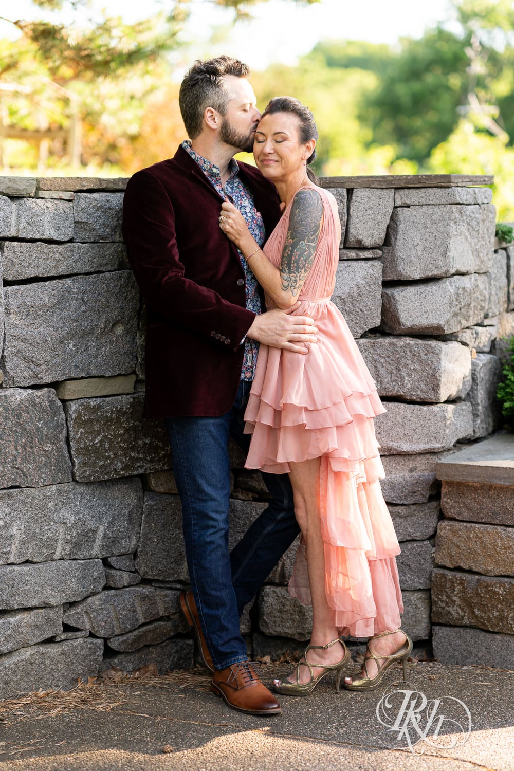 Man in red velvet jacket and a woman in a pink dress kiss in front of a stone wall at the Minnesota Landscape Arboretum in Chaska, Minnesota.