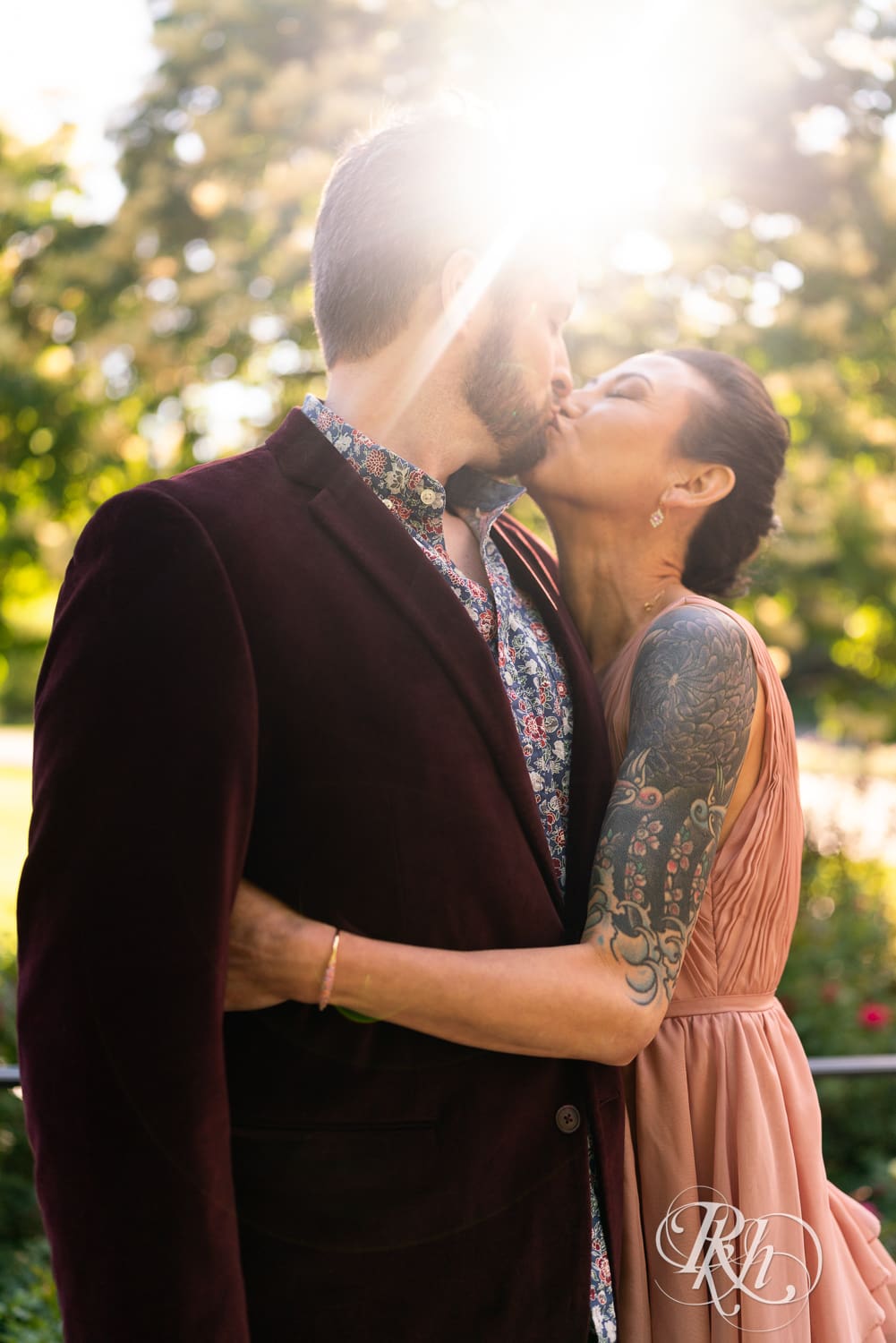 Man in red velvet jacket and a woman in a pink dress kiss in sunlight at the Minnesota Landscape Arboretum in Chaska, Minnesota.