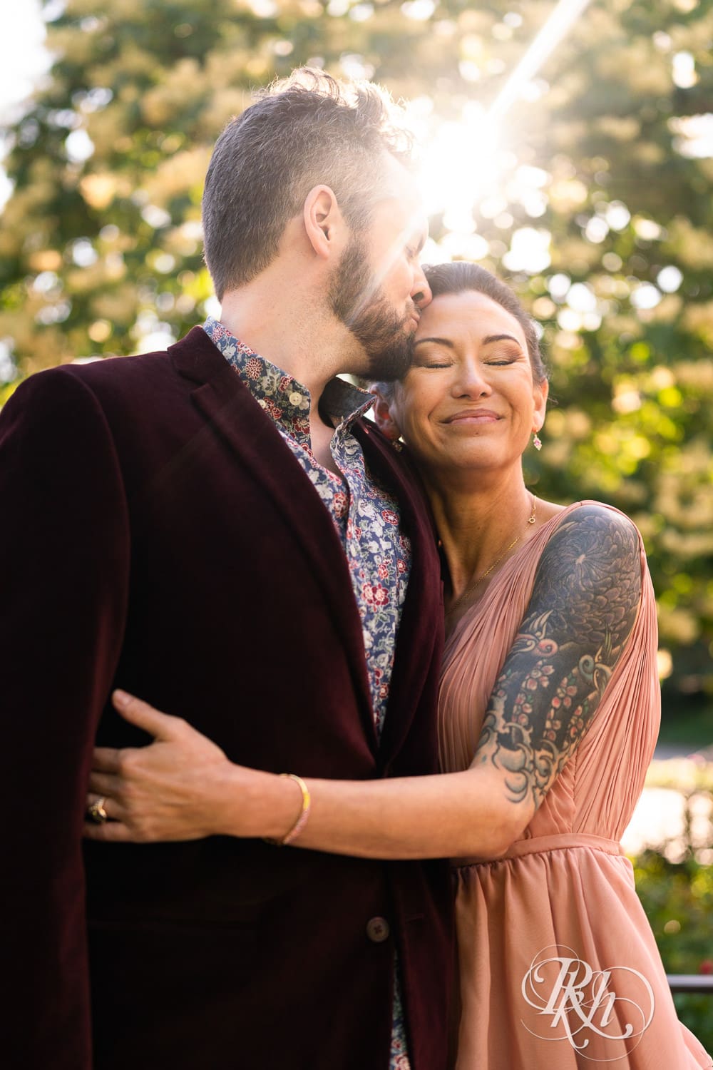 Man in red velvet jacket and a woman in a pink dress kiss in sunlight at the Minnesota Landscape Arboretum in Chaska, Minnesota.