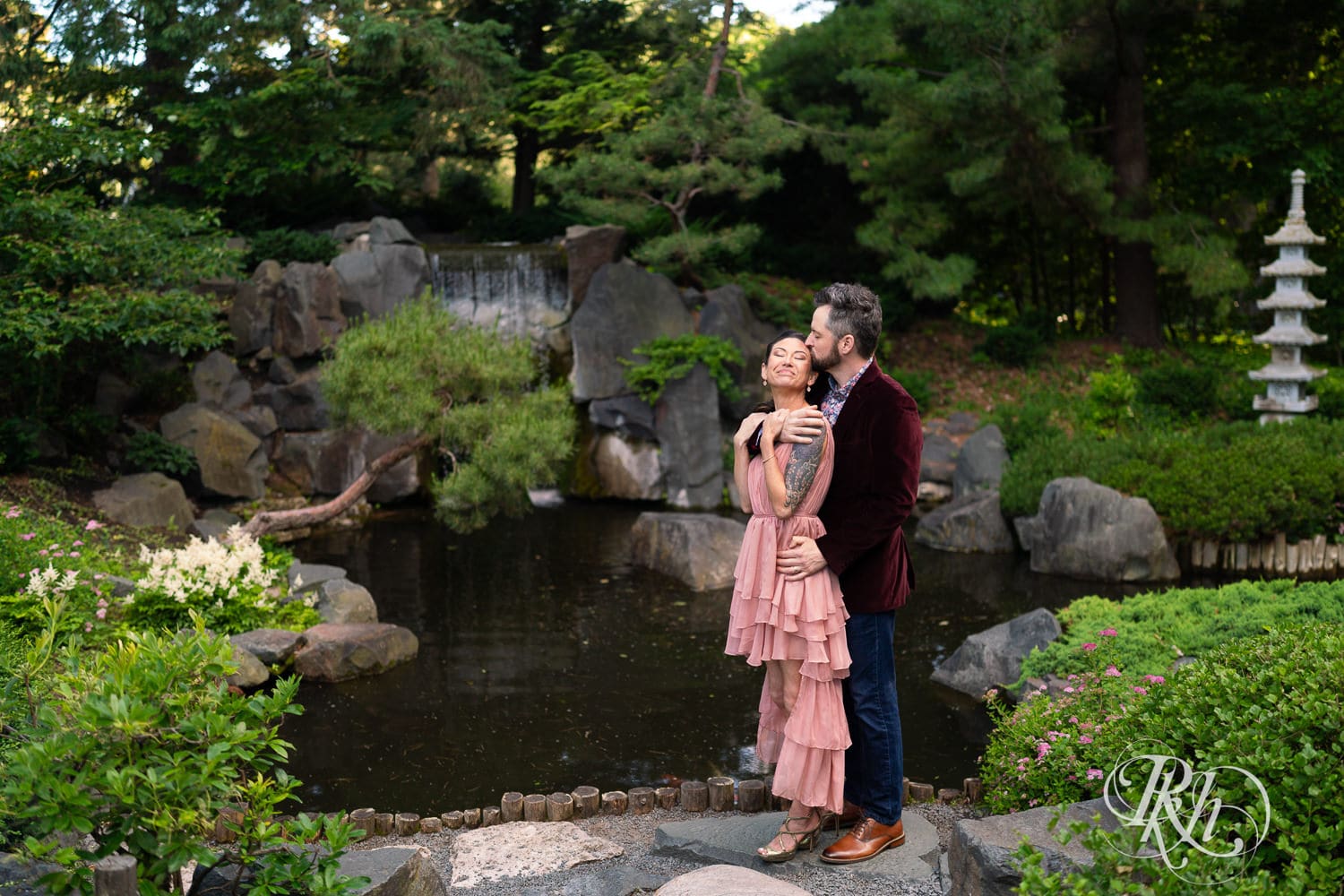 Man in red velvet jacket and a woman in a pink dress kiss in in front of waterfall at Minnesota Landscape Arboretum in Chaska, Minnesota.