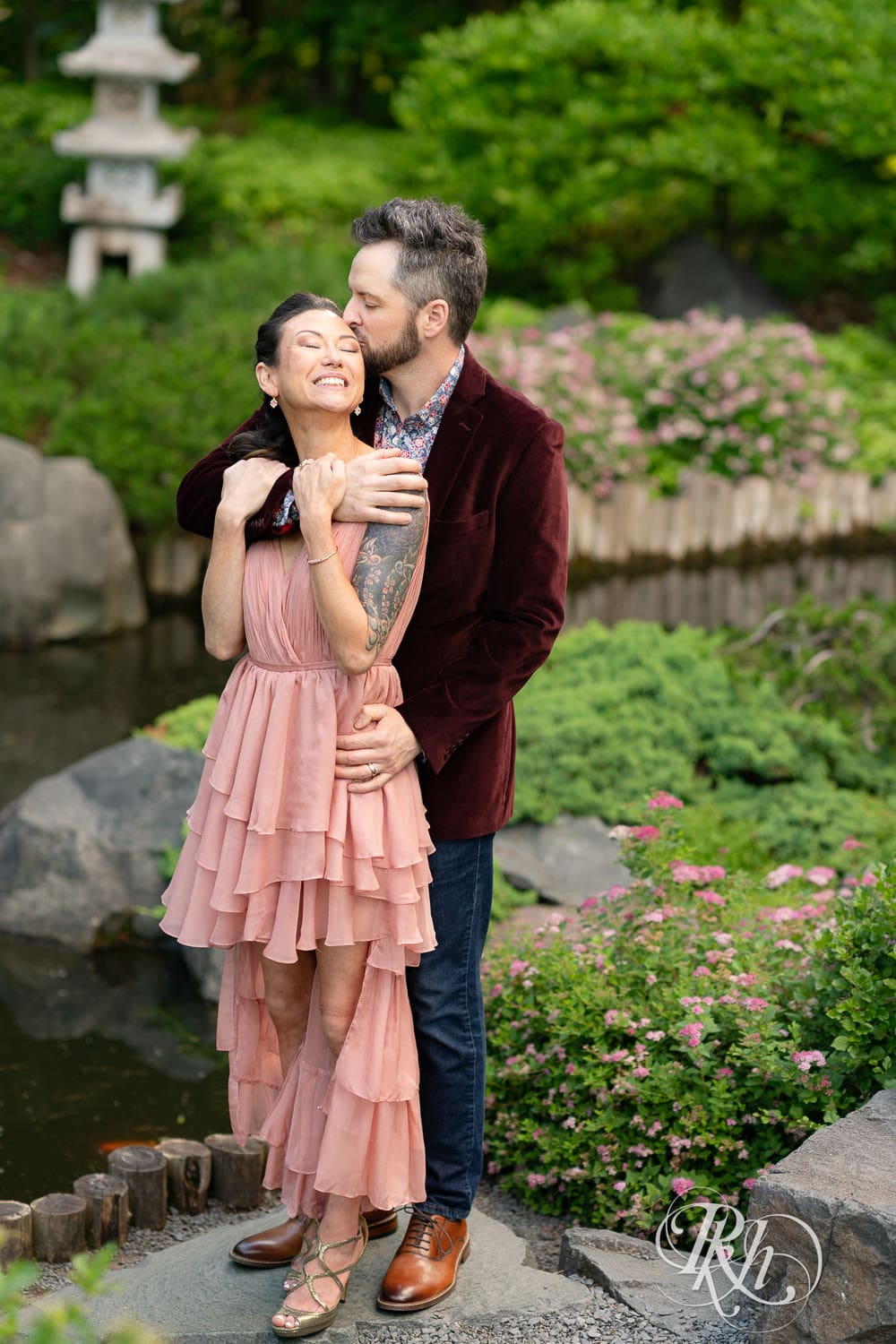 Man in red velvet jacket and a woman in a pink dress kiss in in front of waterfall at Minnesota Landscape Arboretum in Chaska, Minnesota.