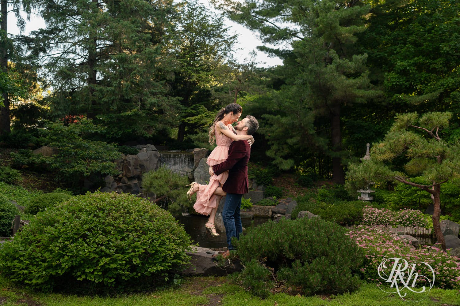 Man in red velvet jacket lifts a woman in a pink dress in front of a waterfall at the Minnesota Landscape Arboretum in Chaska, Minnesota.