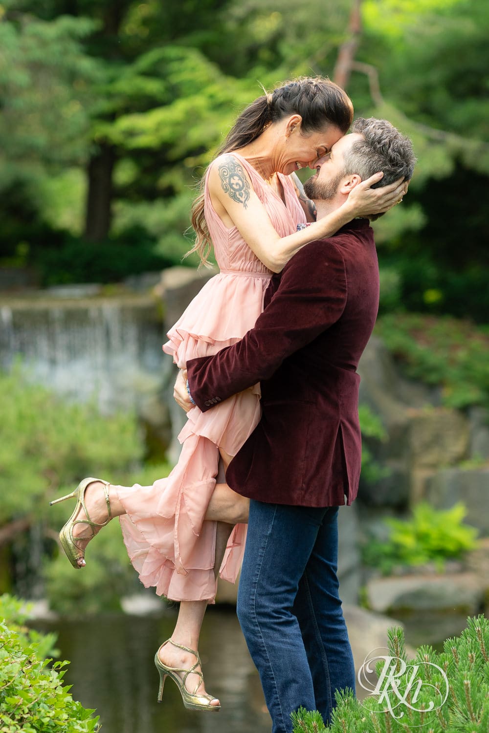 Man in red velvet jacket lifts a woman in a pink dress in front of a waterfall at the Minnesota Landscape Arboretum in Chaska, Minnesota.