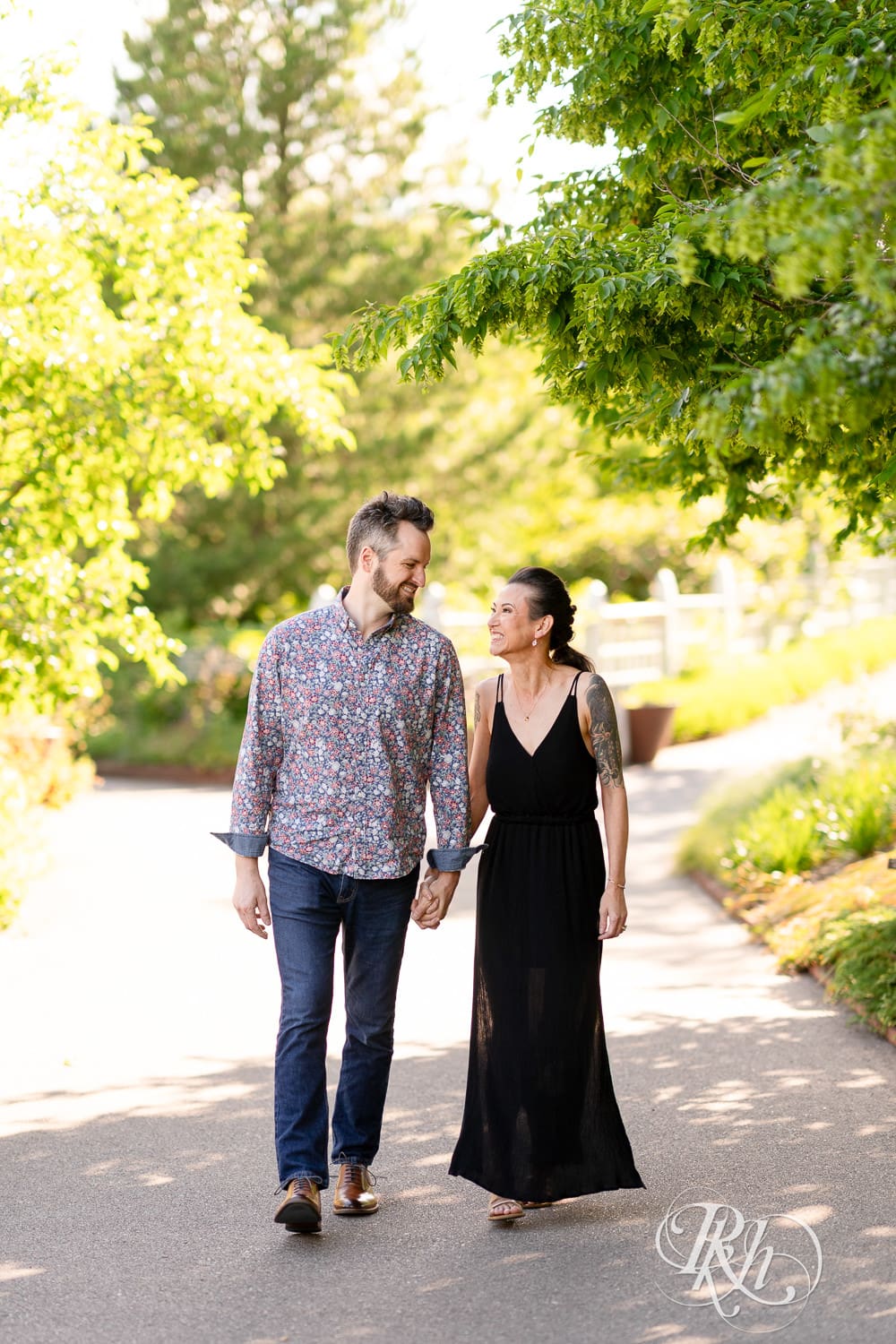 Man and woman in a black dress walk together during sunset at their Minnesota Landscape Arboretum engagement photography session.