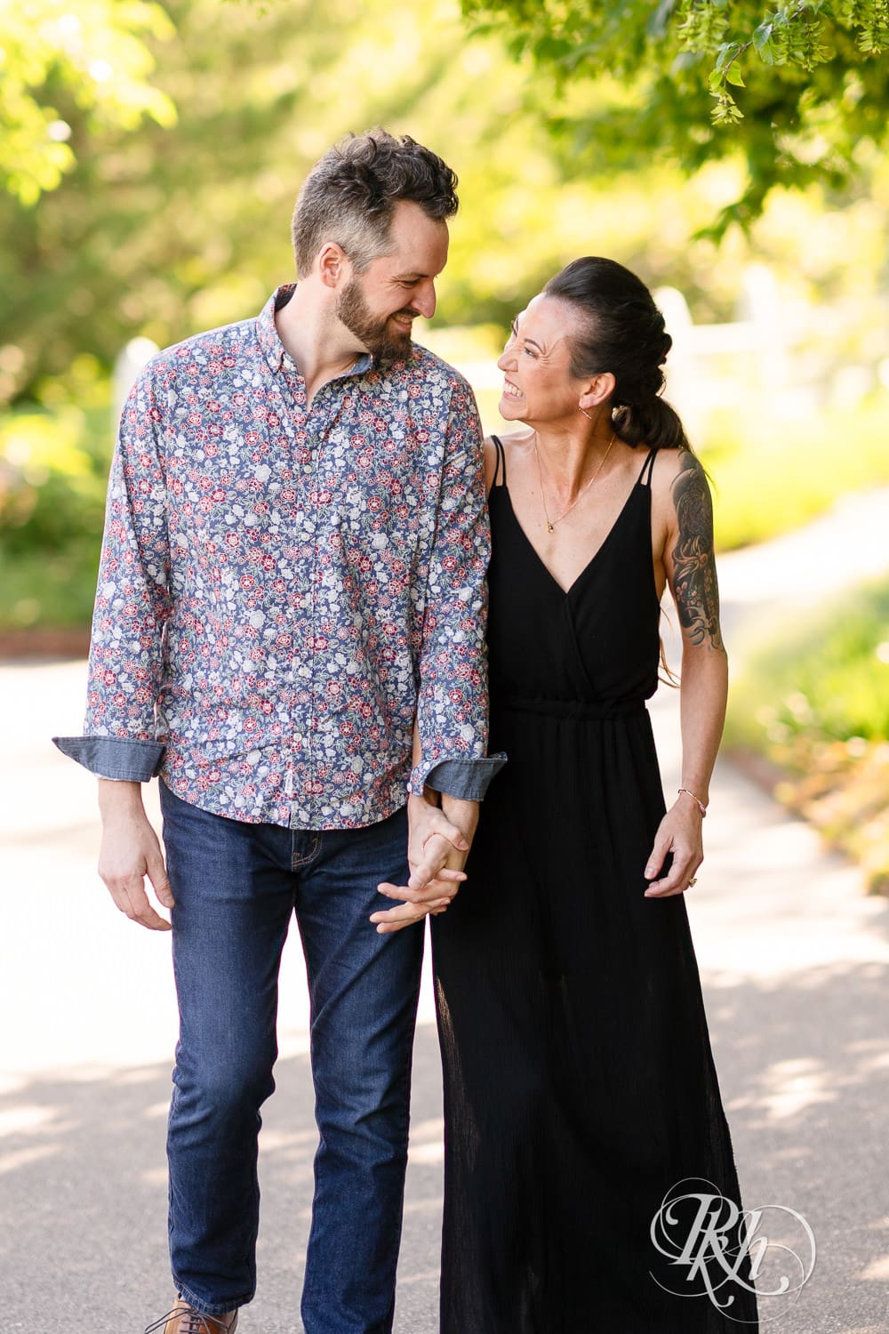 Man and woman in a black dress walk together during sunset at their Minnesota Landscape Arboretum engagement photography session.