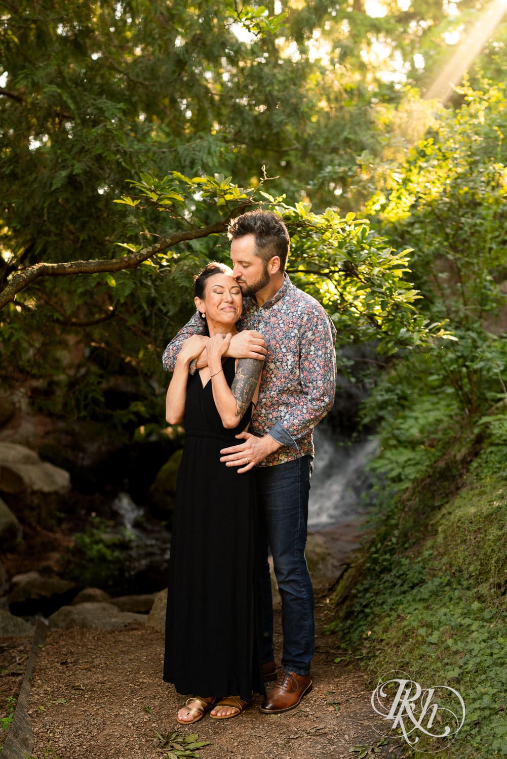 Man and woman in a black dress kiss in the sunlight with trees and waterfall in the background in Chaska, Minnesota.