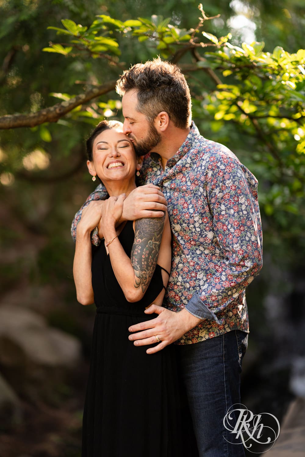 Man and woman in a black dress kiss in the sunlight with trees and waterfall in the background in Chaska, Minnesota.