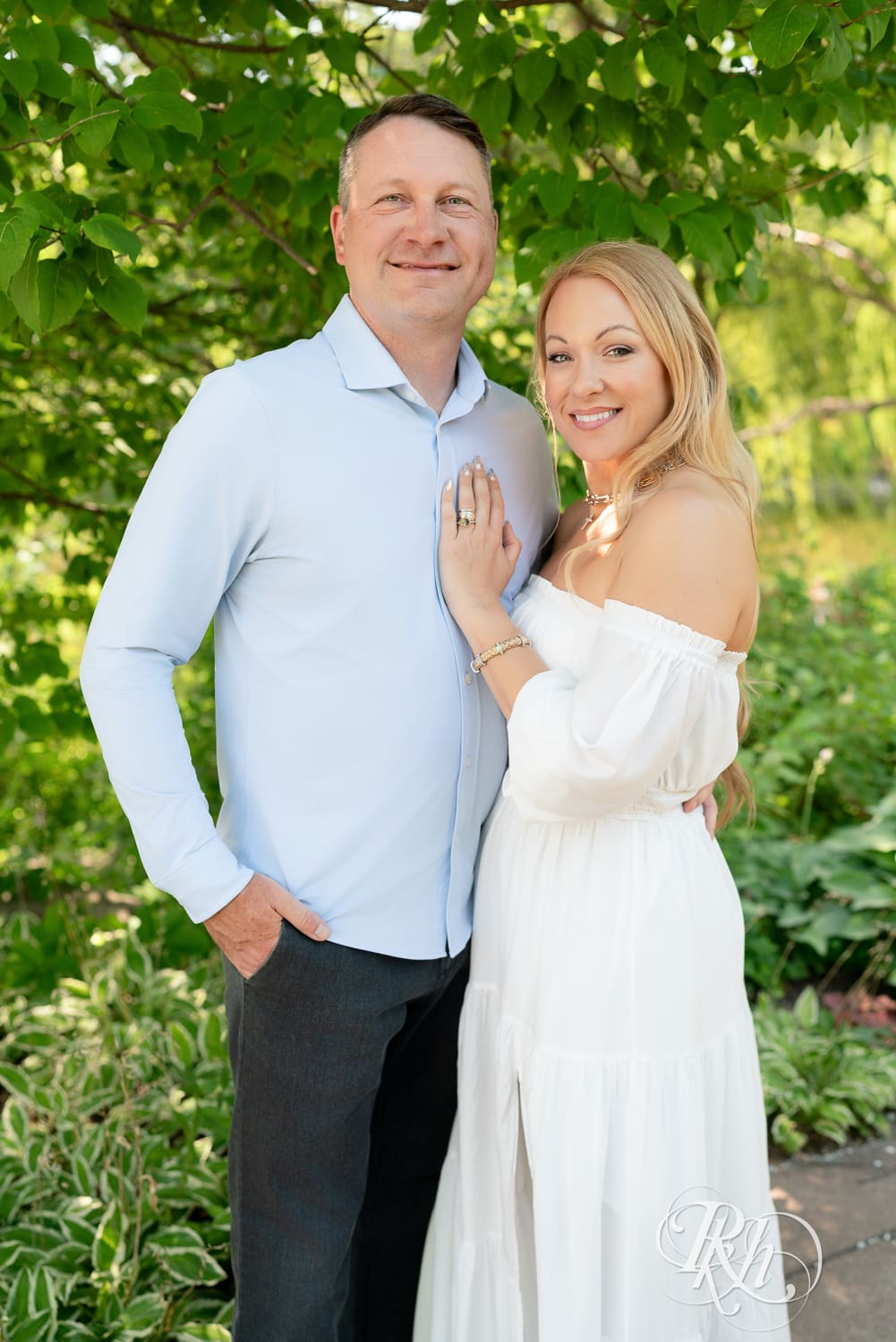 Man in jeans and blond woman in a white dress smile in front of a tree at Centennial Lake Park in Edina, Minnesota.