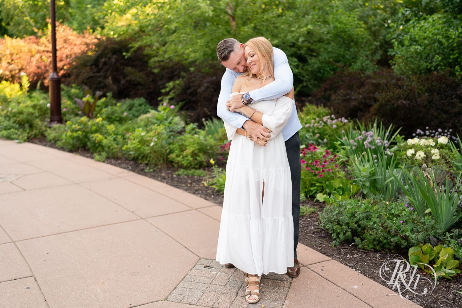 Man in jeans and blond woman in a white dress snuggle in front of flowers at Centennial Lake Park in Edina, Minnesota.