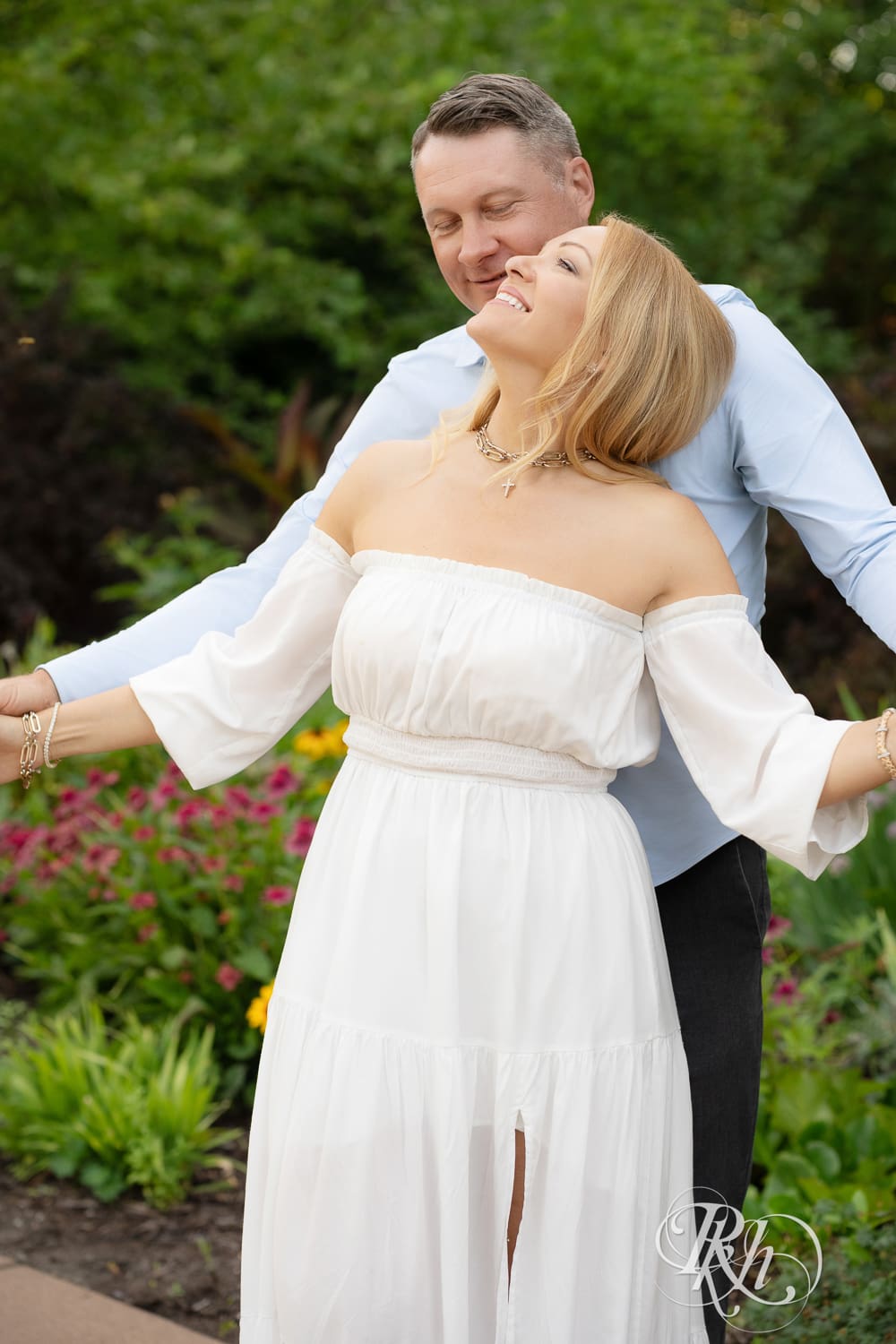 Man in jeans and blond woman in a white dress snuggle in front of flowers at Centennial Lake Park in Edina, Minnesota.