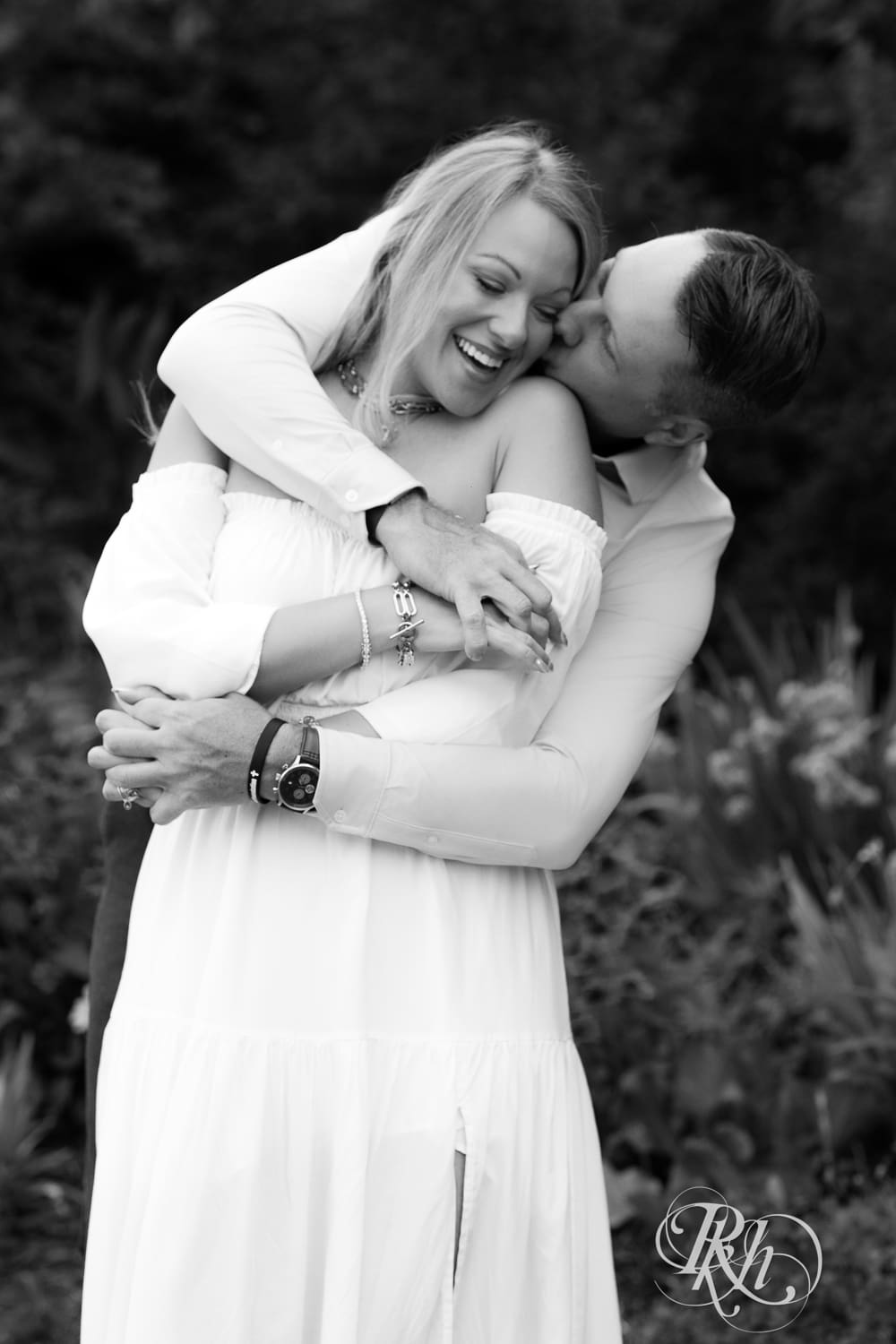 Man in jeans and blond woman in a white dress snuggle in front of flowers at Centennial Lake Park in Edina, Minnesota.