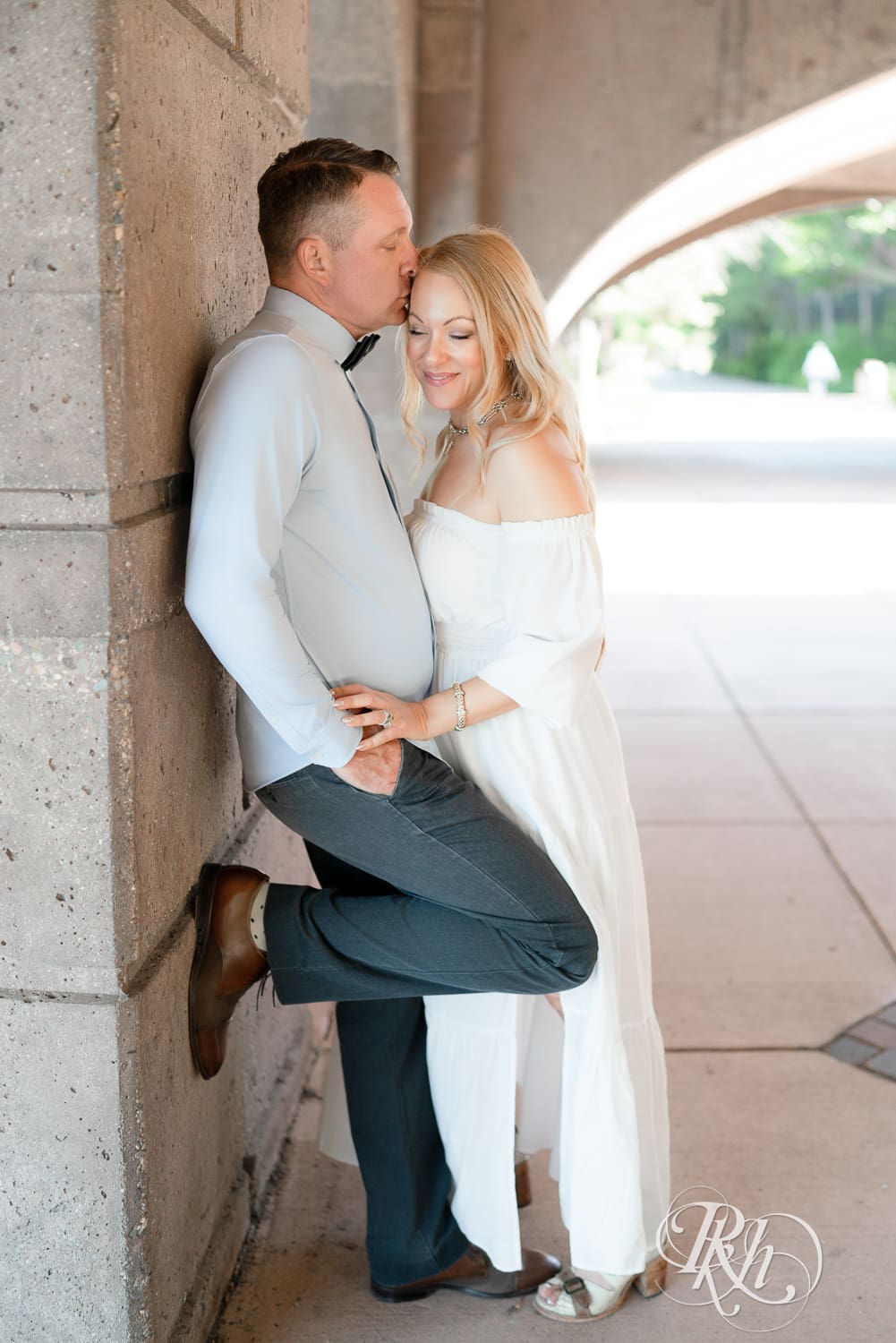Man in jeans and blond woman in a white dress smile under a bridge at Centennial Lake Park in Edina, Minnesota.