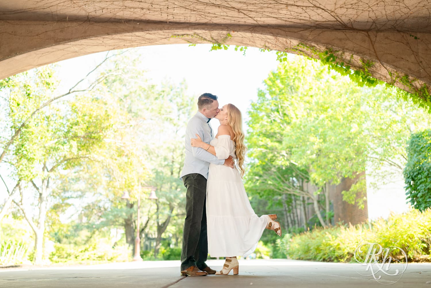 Man in jeans and blond woman in a white dress kiss under a bridge at Centennial Lake Park in Edina, Minnesota.