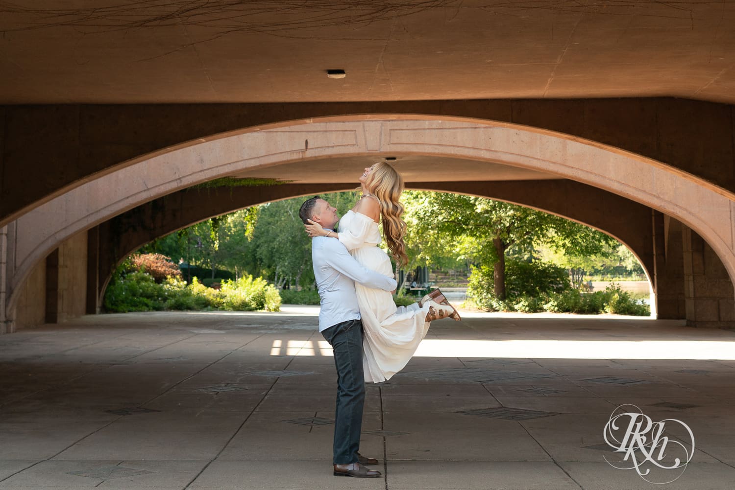 Man in jeans lifts blond woman in a white dress under a bridge at Centennial Lake Park in Edina, Minnesota.