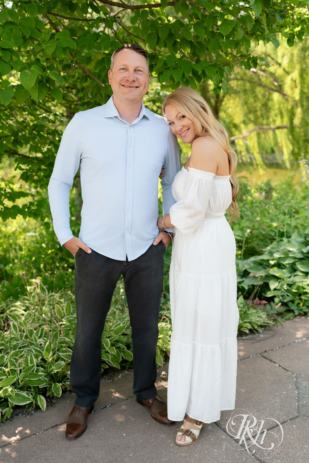 Man in jeans and blond woman in a white dress smile in front of a tree at Centennial Lake Park in Edina, Minnesota.