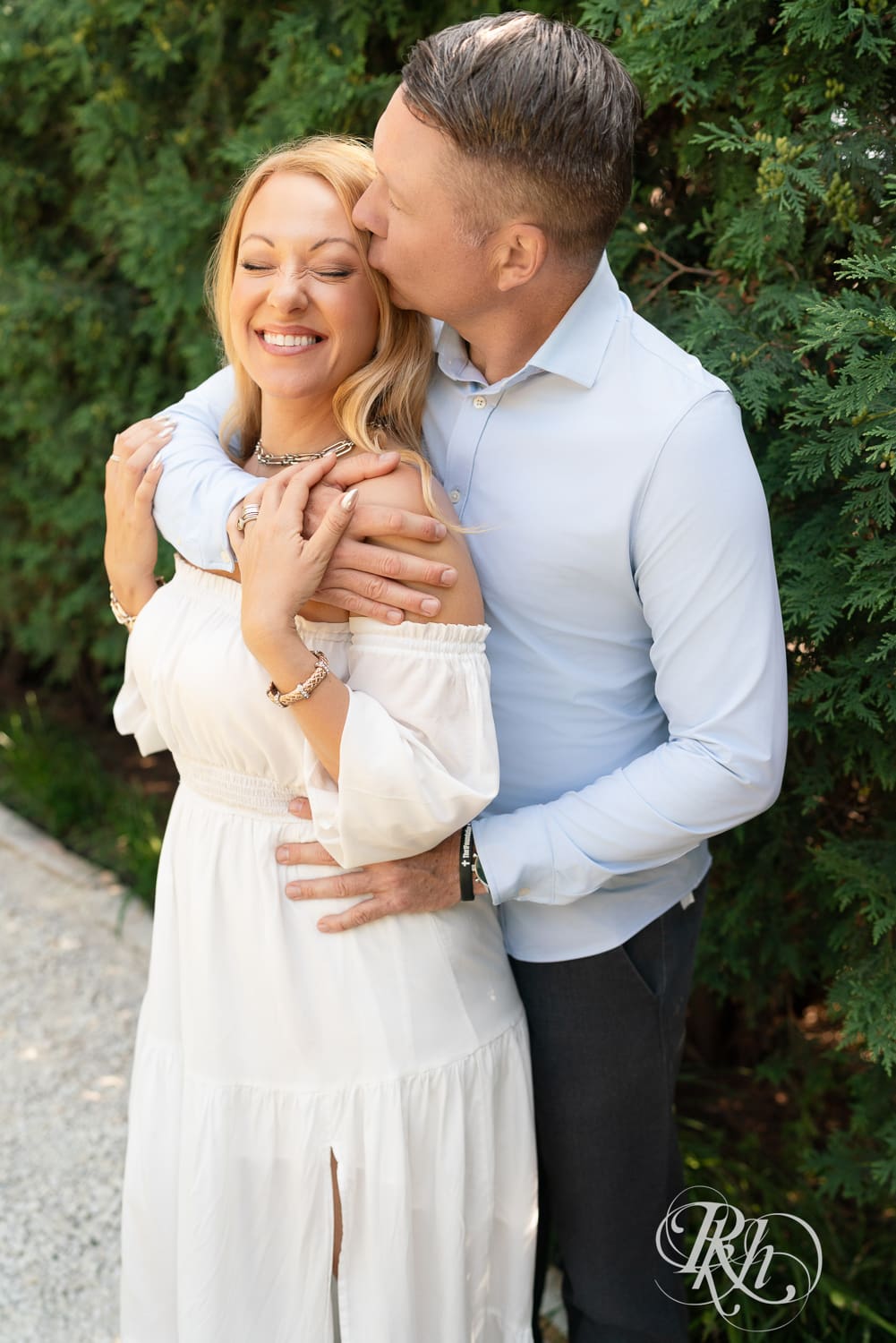 Man in jeans and blond woman in a white dress kiss in front of a tree at Centennial Lake Park in Edina, Minnesota.