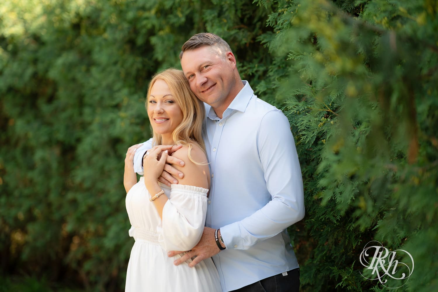 Man in jeans and blond woman in a white dress smile in front of a tree at Centennial Lake Park in Edina, Minnesota.