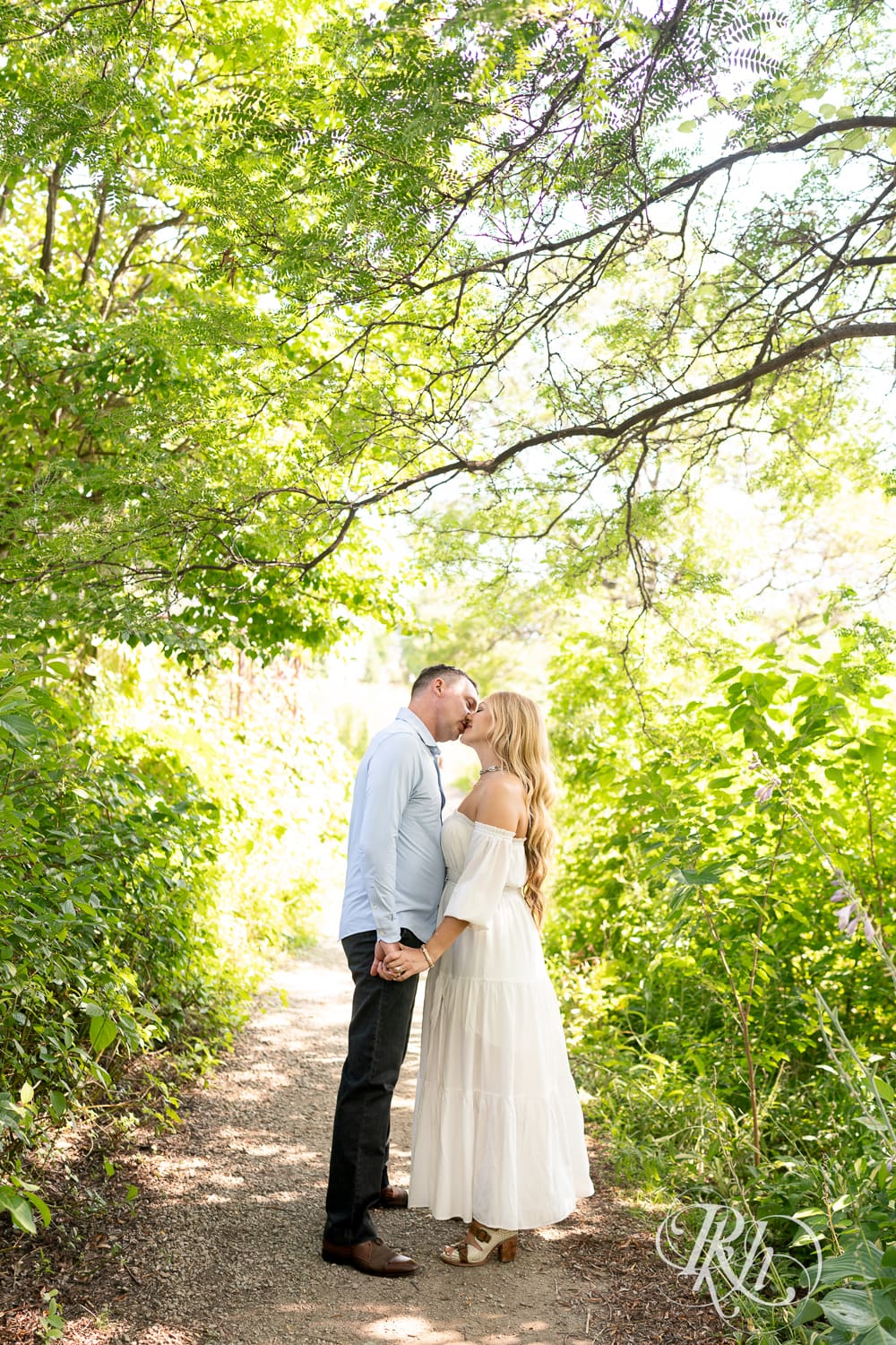 Man in jeans and blond woman in a white dress kiss between trees on a path at Centennial Lake Park in Edina, Minnesota.