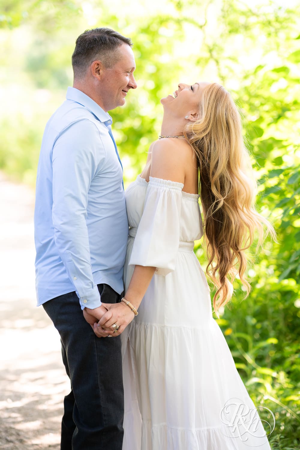 Man in jeans and blond woman in a white dress laugh between trees on a path at Centennial Lake Park in Edina, Minnesota.