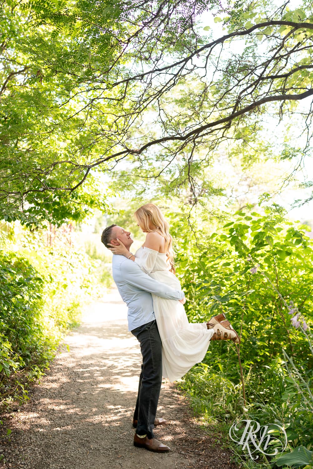 Man in jeans lifts blond woman in a white dress between trees on a path at Centennial Lake Park in Edina, Minnesota.