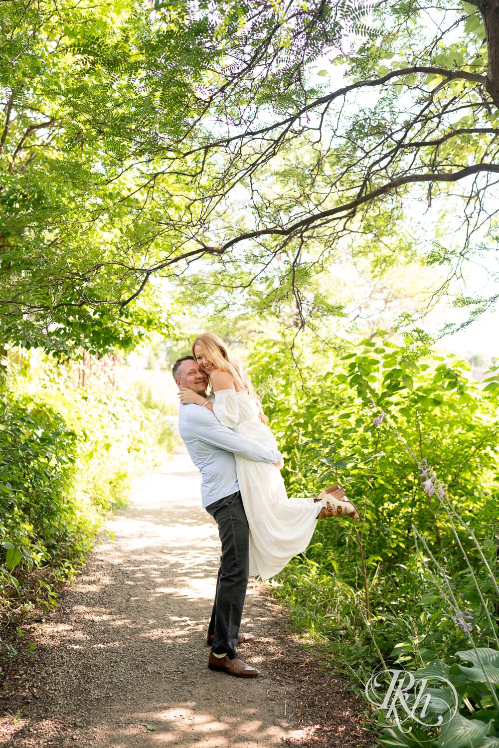 Man in jeans lifts blond woman in a white dress between trees on a path at Centennial Lake Park in Edina, Minnesota.